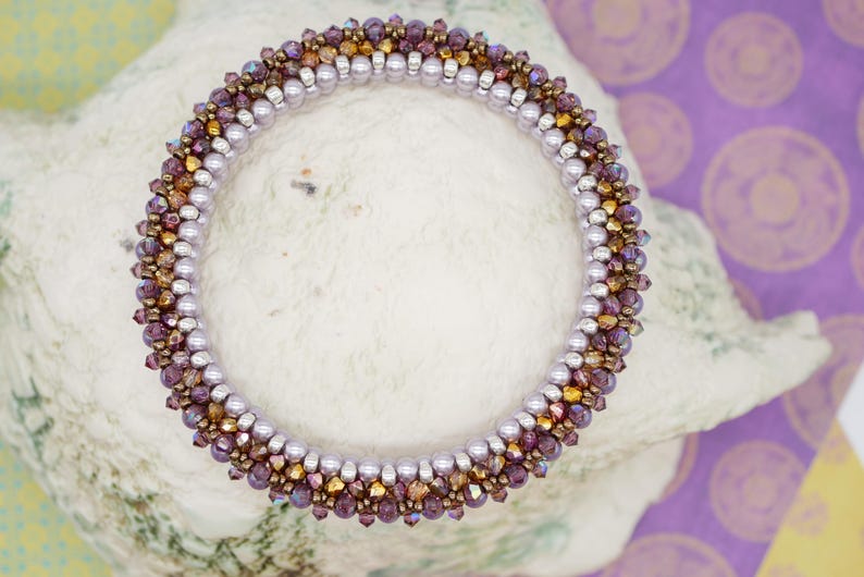 May include: A beaded bracelet with a gradient of purple, gold, and silver beads. The bracelet is made with small, round beads and is arranged in a spiral pattern.