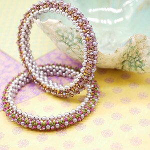 May include: Two beaded bracelets with a mix of purple, pink, and gold beads. The bracelets are stacked on top of each other and are lying on a patterned surface.