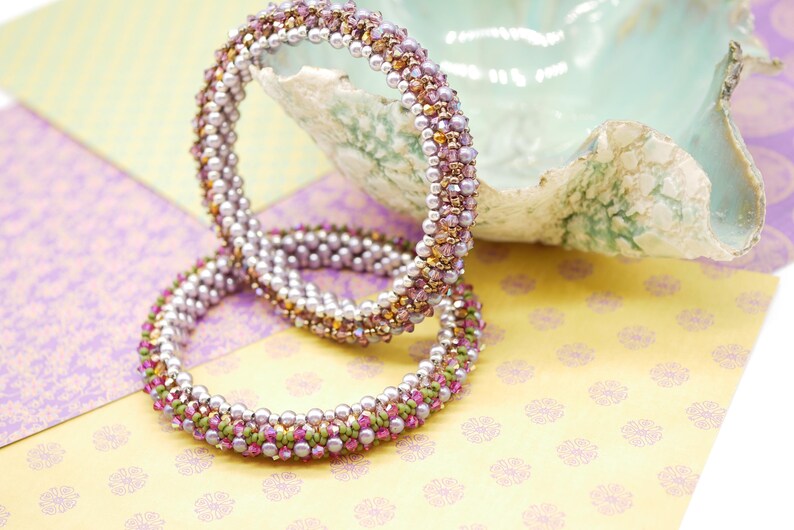 May include: Two beaded bracelets with a mix of purple, pink, and gold beads. The bracelets are stacked on top of each other and are lying on a patterned surface.