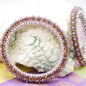 May include: Two beaded bracelets with a purple, pink, and gold color scheme. The bracelets are made with small, round beads and are stacked on top of each other.