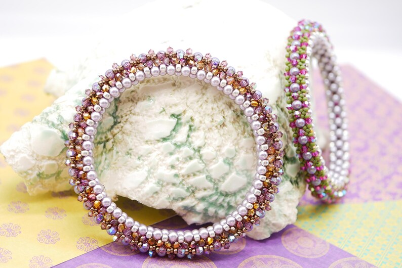 May include: Two beaded bracelets with a purple, pink, and gold color scheme. The bracelets are made with small, round beads and are stacked on top of each other.