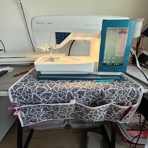 May include: A white Husqvarna Viking Designer Sapphire 85 sewing machine with a teal touchscreen display. The machine sits on a table covered with a gray and white heart-patterned cover. Sewing supplies are visible.