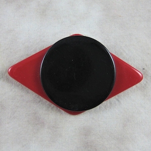 May include: A black and red plastic brooch in the shape of a diamond with a black circle in the center.