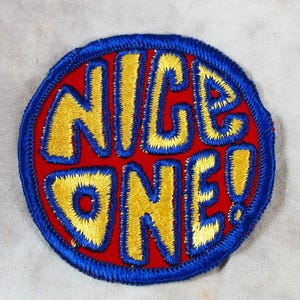 May include: A round embroidered patch with the words "NICE ONE!" in yellow, outlined in blue, on a red background. The patch has a blue border and is likely for clothing or accessories.