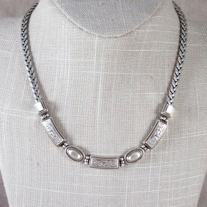 May include: A silver necklace and matching square earrings set. The necklace has a braided chain and rectangular and oval decorative elements. The earrings have a matching design. The jewellery is displayed on a mannequin.