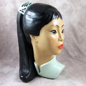 May include: Vintage Asian-inspired head bust with black hair in a ponytail. The bust has a light skin tone, red lipstick, and a decorative white and green hair accessory. The neck is a light green color.