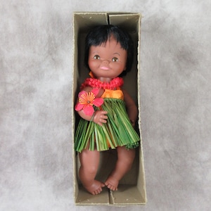 May include: A vintage doll in a cardboard box. The doll has dark hair, brown skin, and is wearing a grass skirt, a flower lei, and holding a pink flower. The doll is a Hawaiian-themed collectible.