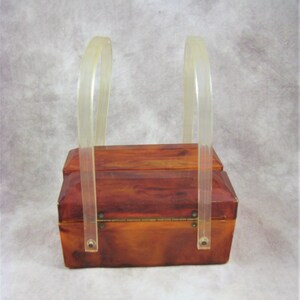 May include: A vintage, rectangular, brown, plastic box with a hinged lid and two clear plastic handles. The box has a wood-grain pattern.