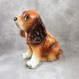 May include: A brown and white ceramic figurine of a sitting cocker spaniel dog. The dog has brown fur with white markings on its chest and paws. The figurine is in a sitting position with its head tilted to the side.