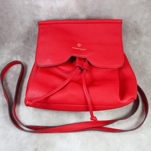 May include: A red leather backpack with a drawstring closure and a small gold logo that says "Vanessa Bruno." The backpack has a long adjustable strap.