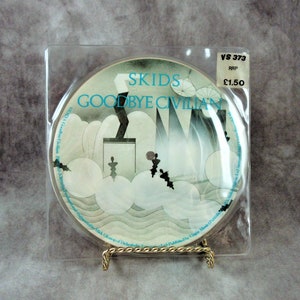 May include: A vinyl record in a clear plastic sleeve with a white label. The label has a blue and white graphic design with the text "SKIDS GOODBYE CIVILIAN" and the record number "VS 373". A price tag is attached to the sleeve with the text "RRP £1.50".