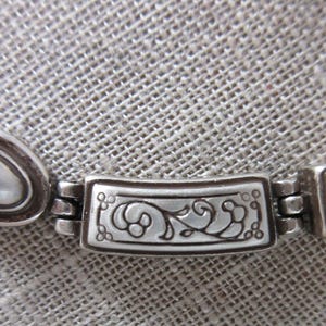 May include: Close-up of a silver necklace. The necklace has oval and rectangular links, with an ornate floral design on the central link. The chain has a braided design. The jewellery is displayed on a textured, grey surface.