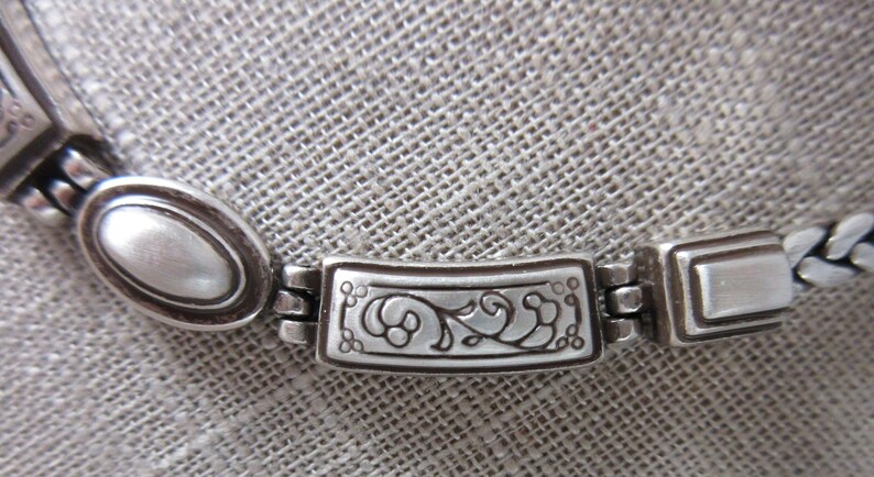 May include: Close-up of a silver necklace. The necklace has oval and rectangular links, with an ornate floral design on the central link. The chain has a braided design. The jewellery is displayed on a textured, grey surface.