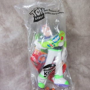 K&ouml;nnte beinhalten: Eine Buzz Lightyear-Actionfigur aus dem Film "Toy Story" in einer durchsichtigen Plastikt&uuml;te. Die Figur ist wei&szlig;, gr&uuml;n und lila, mit dem Burger King-Logo auf der Verpackung. Die T&uuml;te hat auch das "Toy Story"-Logo.