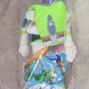 K&ouml;nnte beinhalten: Eine Buzz Lightyear-Actionfigur in einer durchsichtigen Plastikt&uuml;te. Die Figur ist &uuml;berwiegend wei&szlig; mit gr&uuml;nen und lila Akzenten und einem gr&uuml;nen Rucksack. Ein Pappschild mit Toy Story-Charakteren ist sichtbar.