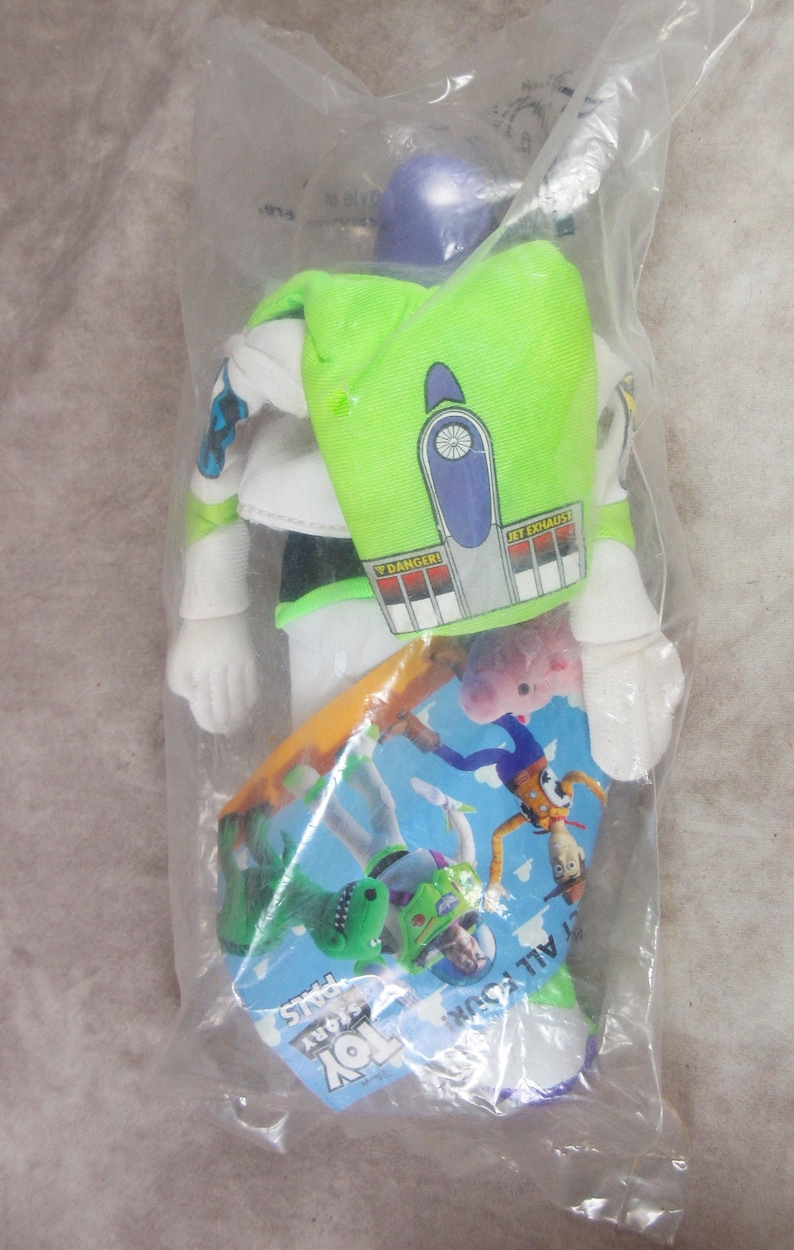 K&ouml;nnte beinhalten: Eine Buzz Lightyear-Actionfigur in einer durchsichtigen Plastikt&uuml;te. Die Figur ist &uuml;berwiegend wei&szlig; mit gr&uuml;nen und lila Akzenten und einem gr&uuml;nen Rucksack. Ein Pappschild mit Toy Story-Charakteren ist sichtbar.