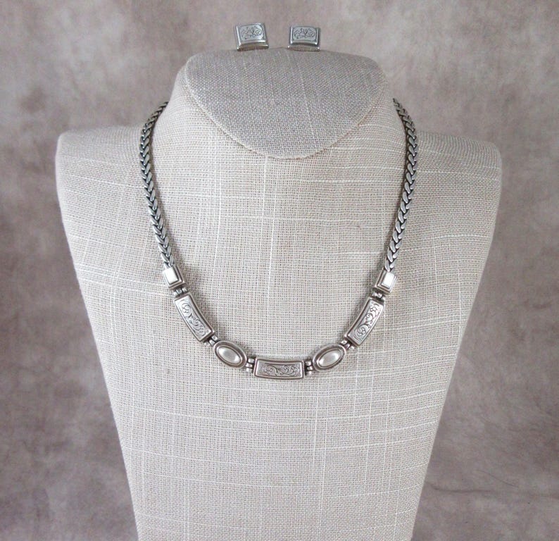 May include: A silver necklace and matching square earrings set. The necklace has a braided chain and rectangular and oval decorative elements. The earrings have a matching design. The jewellery is displayed on a mannequin.