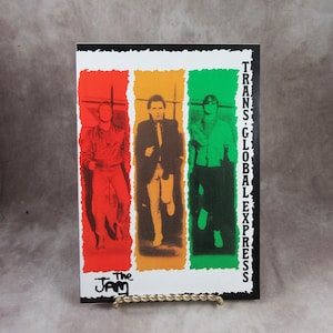 May include: A framed print featuring three figures in colorful vertical panels. The figures are in red, orange, and green. The text "TRANS-GLOBAL EXPRESS" is on the right side, and "The Jam" is at the bottom.