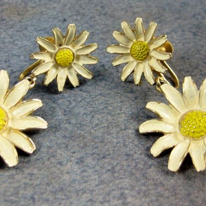 May include: A pair of gold tone clip-on earrings featuring white enamel daisy flowers with yellow centers.