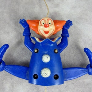 May include: A blue and orange plastic clown toy with a pull string. The clown has a big smile and is wearing a blue and white costume. The clown's arms are raised in the air and its legs are spread out.