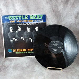 May include: A black vinyl record with a black and white album cover. The album cover features a band of four men, with the title "The Beetle Beat" and the song title "I Want To Hold Your Hand/The Buggs".