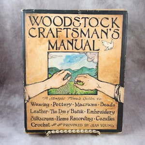 May include: A vintage "Woodstock Craftsman's Manual" book cover. The cover is tan with black lettering and illustrations. The title is at the top, with a drawing of hands crafting in the center. The book lists crafts like weaving, pottery, and macrame.