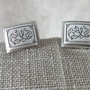 May include: A pair of square silver cufflinks. Each cufflink features a decorative floral design within a rectangular frame. The design is detailed with curved lines and small circular accents, set against a neutral background.