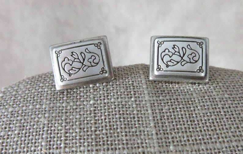 May include: A pair of square silver cufflinks. Each cufflink features a decorative floral design within a rectangular frame. The design is detailed with curved lines and small circular accents, set against a neutral background.