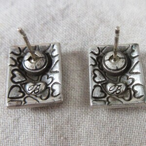 May include: A pair of silver stud earrings. Each rectangular earring features a floral and heart design, with the letter "B" inscribed. The earrings have a post and a round back.