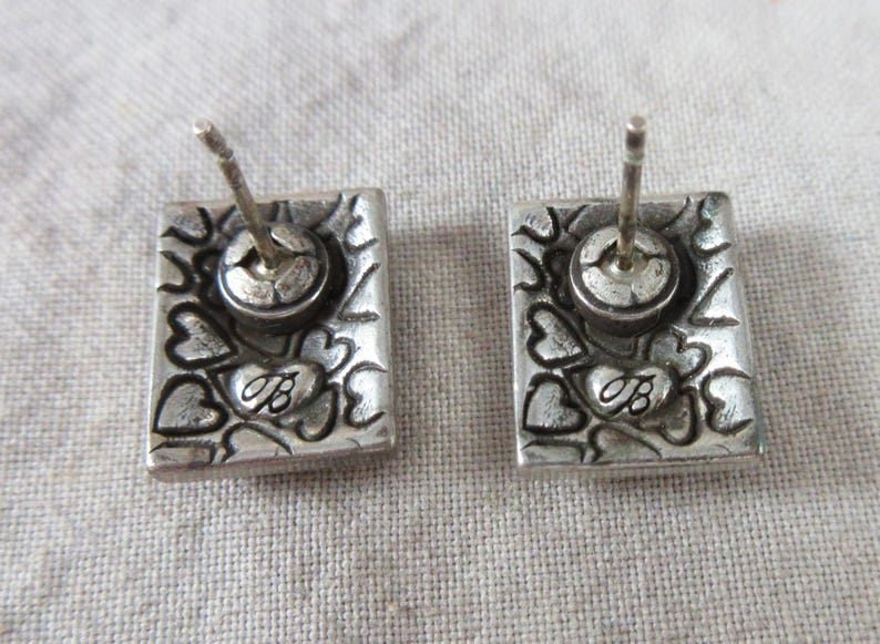 May include: A pair of silver stud earrings. Each rectangular earring features a floral and heart design, with the letter "B" inscribed. The earrings have a post and a round back.