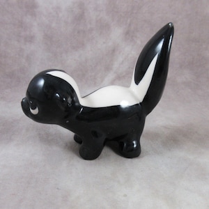May include: A black and white ceramic figurine of a skunk. The skunk is standing on all fours with its tail raised. The figurine is about 4 inches tall.