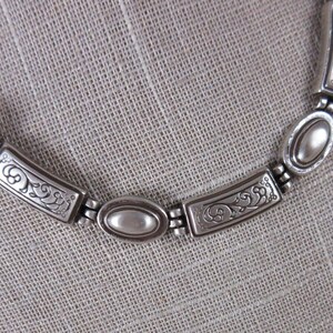 May include: Silver necklace featuring a braided chain and decorative rectangular and oval links. The rectangular links have an ornate, etched design. The necklace is displayed on a neutral-coloured fabric.