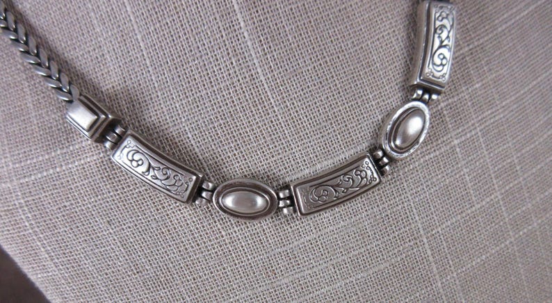 May include: Silver necklace featuring a braided chain and decorative rectangular and oval links. The rectangular links have an ornate, etched design. The necklace is displayed on a neutral-coloured fabric.