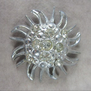 May include: A silver sunburst brooch with clear rhinestones.