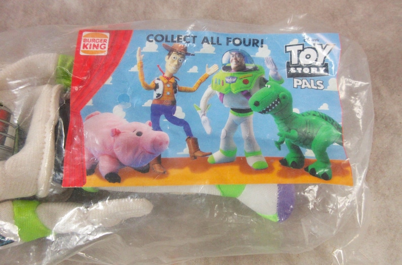 K&ouml;nnte beinhalten: Ein versiegelter Plastikbeutel mit einem Werbeartikel von Burger King. Der Artikel zeigt eine farbenfrohe Karte mit den Toy Story-Charakteren Woody, Buzz Lightyear, Rex und Hamm. Auf der Karte steht "ALLE VIER SAMMELN!" und "TOY STORY PALS".