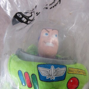 K&ouml;nnte beinhalten: Eine Buzz Lightyear-Actionfigur in einer durchsichtigen Plastikt&uuml;te. Die Figur hat einen gr&uuml;nen und wei&szlig;en Raumanzug mit einem blau-wei&szlig;en Emblem. Das Gesicht ist hellrosa mit blauen Augen und einem strengen Ausdruck. Die T&uuml;te hat Text.
