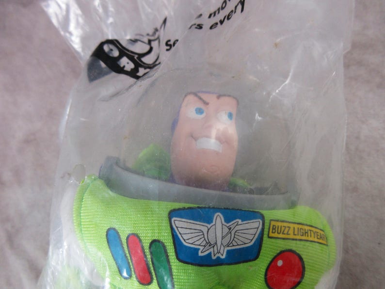 K&ouml;nnte beinhalten: Eine Buzz Lightyear-Actionfigur in einer durchsichtigen Plastikt&uuml;te. Die Figur hat einen gr&uuml;nen und wei&szlig;en Raumanzug mit einem blau-wei&szlig;en Emblem. Das Gesicht ist hellrosa mit blauen Augen und einem strengen Ausdruck. Die T&uuml;te hat Text.