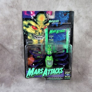 May include: A green and purple Mars Attacks action figure of a spider-like alien with a clear dome head. The figure is packaged in a plastic blister pack with a green and black card back. The card back features the Mars Attacks logo and the text "Software Included".