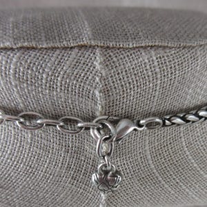 May include: Silver necklace with a woven chain design. The necklace has a lobster clasp and a small charm. The necklace is displayed on a neutral-coloured fabric.