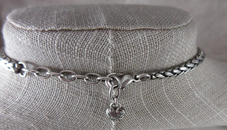 May include: Silver necklace with a woven chain design. The necklace has a lobster clasp and a small charm. The necklace is displayed on a neutral-coloured fabric.
