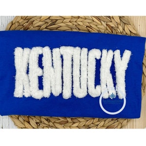 May include: A royal blue t-shirt with the word "KENTUCKY" in white fluffy letters. The letters are raised and have a fuzzy texture.