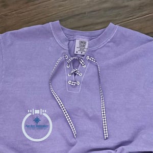 May include: A lavender Comfort Colors sweatshirt with a lace-up neckline. The lace-up detail features a white and black gingham ribbon. The sweatshirt has a crew neck and a tag that reads "Comfort Colors". The logo for "Just Sew Embroidery" is visible.