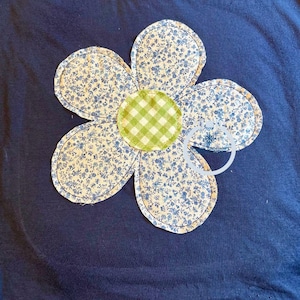 May include: Navy blue t-shirt featuring a large, appliqued flower. The flower has five petals with a blue floral pattern and a green gingham center. The petals are outlined with a blue stitch.