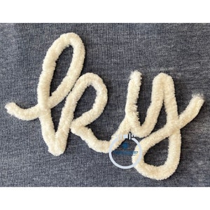 May include: A close-up of the word "Hey" embroidered in white fluffy yarn on a dark blue fabric.