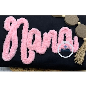 Könnte beinhalten: Rosa flauschige Buchstaben, die "mama" auf einem schwarzen Stoffhintergrund buchstabieren.