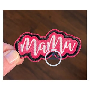 May include: Pink and black embroidered patch with the word "mama" in white cursive lettering.