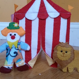 Può includere: Una tenda da circo in feltro rosso e bianco a strisce con una patta in feltro giallo. Una bambola di clown con il naso rosso e i capelli arancioni si trova davanti alla tenda. Una bambola di leone in feltro marrone è seduta davanti alla tenda.