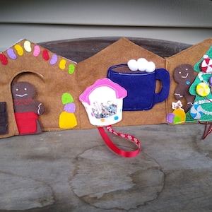 May include: A felt Christmas-themed decoration featuring a gingerbread house, a blue mug with marshmallows, and a Christmas tree. The gingerbread house has a gingerbread man and candy decorations. A cupcake-shaped ornament hangs in front.