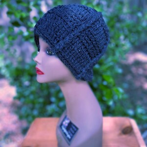 May include: A dark gray, hand-knitted hat with a textured pattern. The hat is displayed on a mannequin head with red lipstick and a neutral skin tone. The background is a blurred view of green foliage.