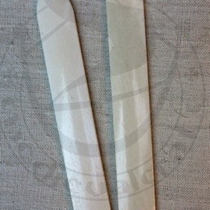 May include: Two off-white bone folders with smooth, elongated bodies and pointed tips. These tools feature a subtle, light-colored pattern across their surfaces, with one folder slightly shorter than the other, both displaying a polished finish.